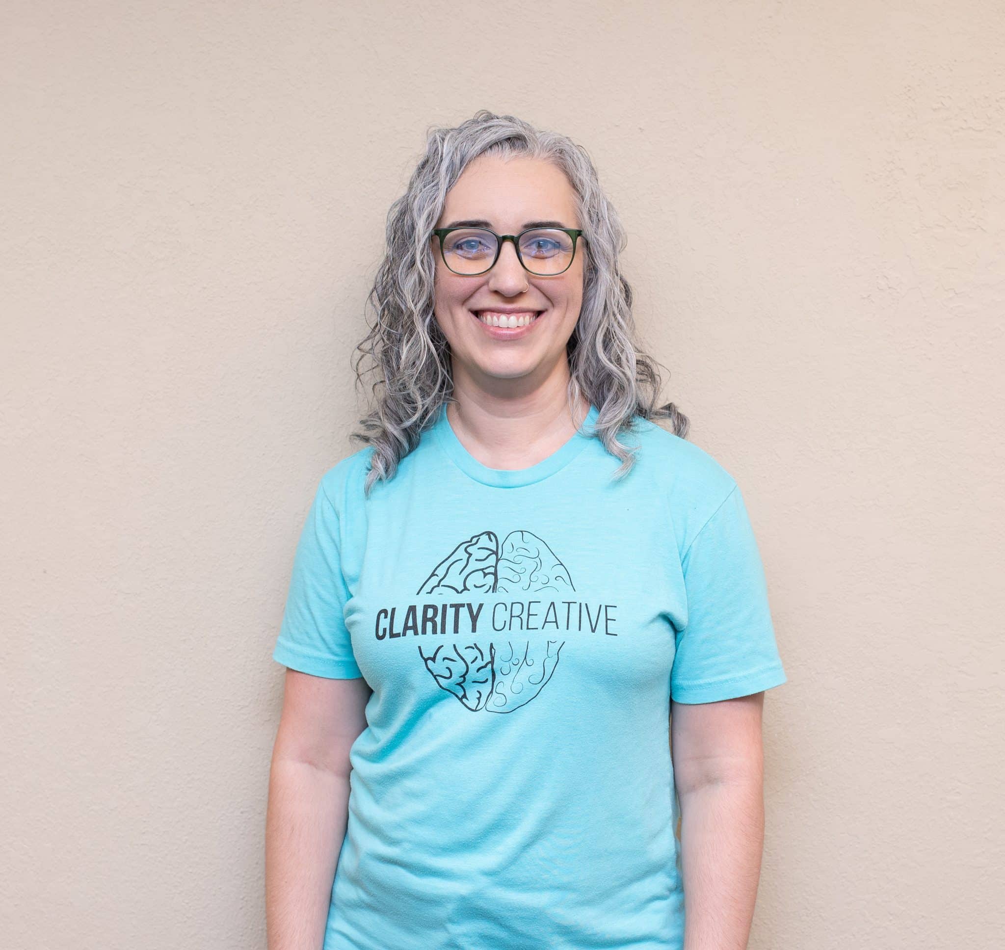 Julie of Clarity Creative Group. She is smiling, wearing glasses, and has curly salt and pepper hair. She is also wearing a teal colored Clarity Creative shirt