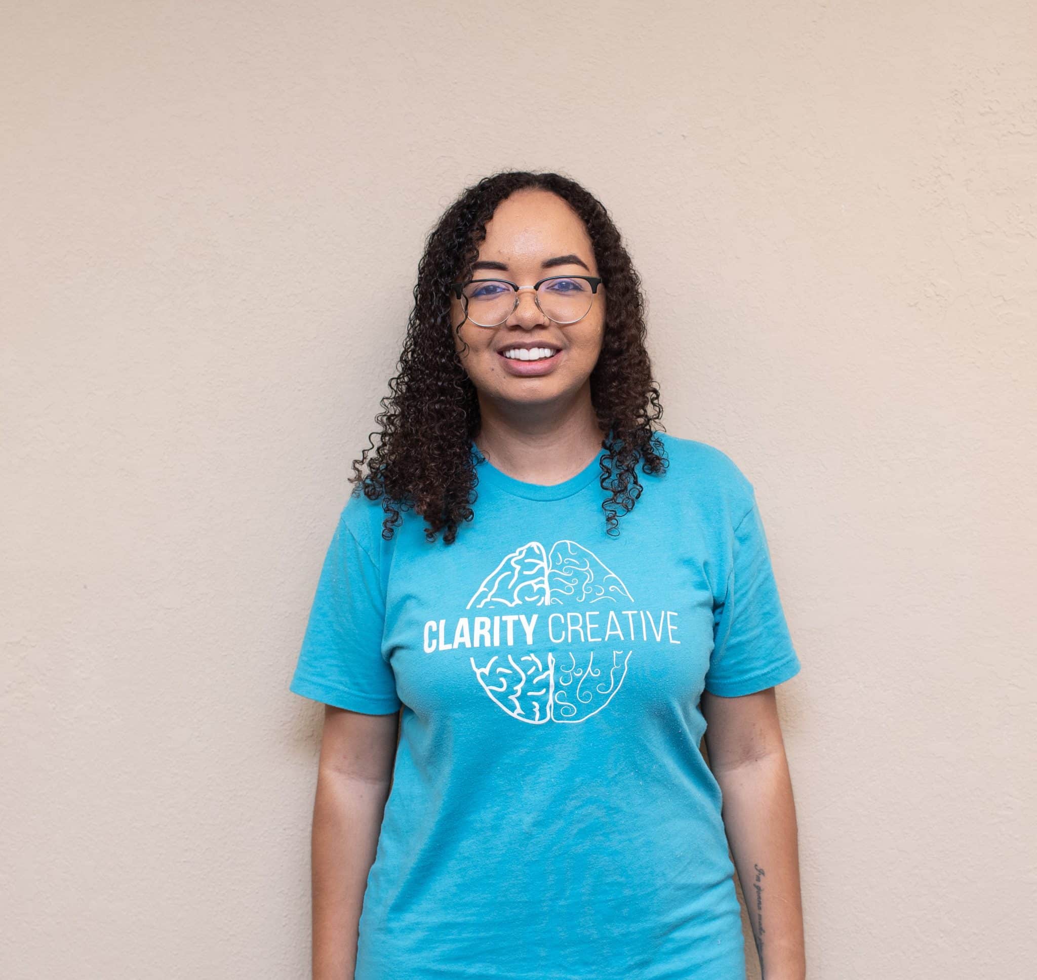 Lux from Clarity Creative Group. She has dark curly hair, glasses, and is wearing a teal Clarity Creative shirt