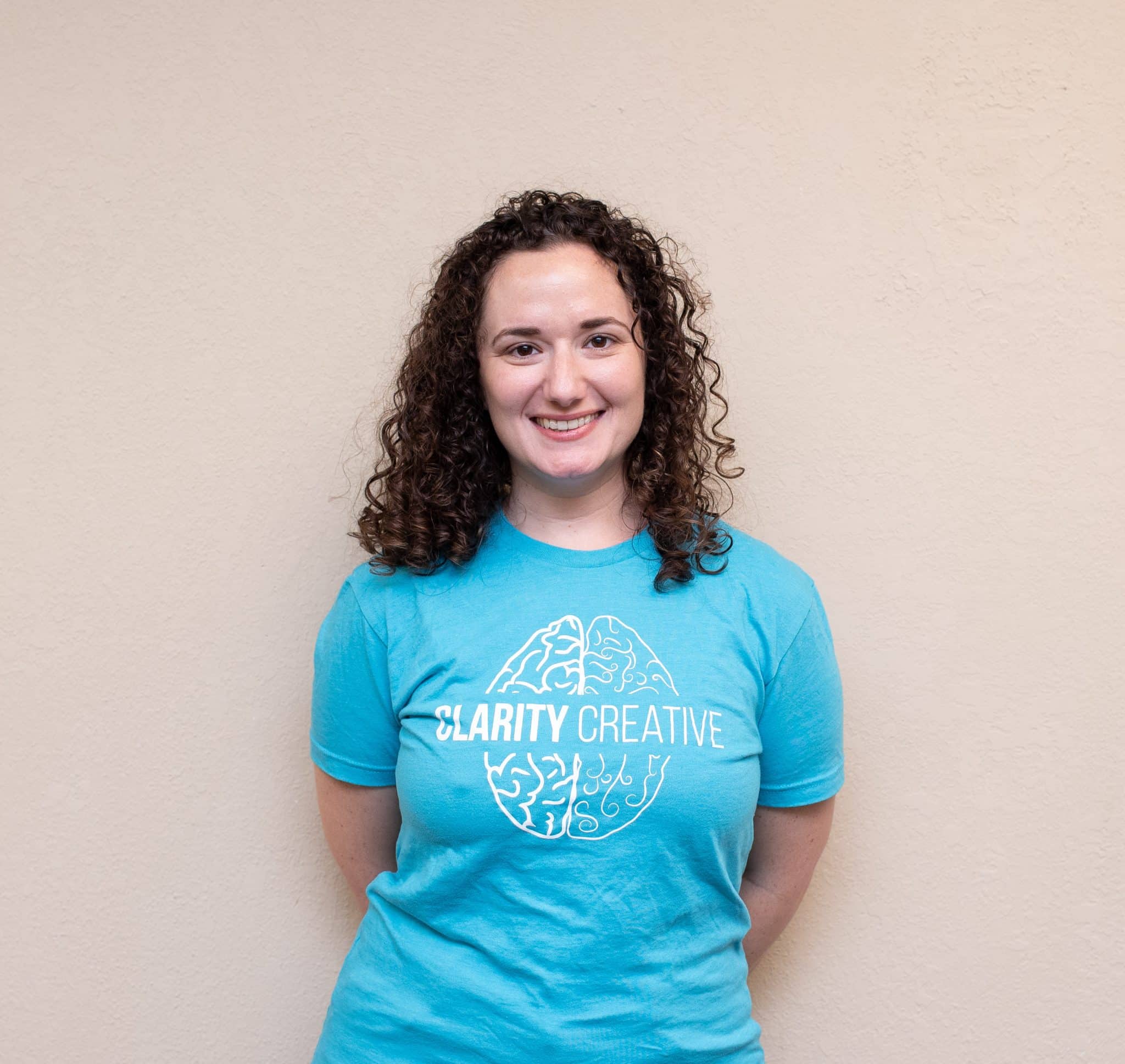 Neri of Clarity Creative Group. She is smiling at the camera, has brown curly hair, and is wearing a teal Clarity shirt.