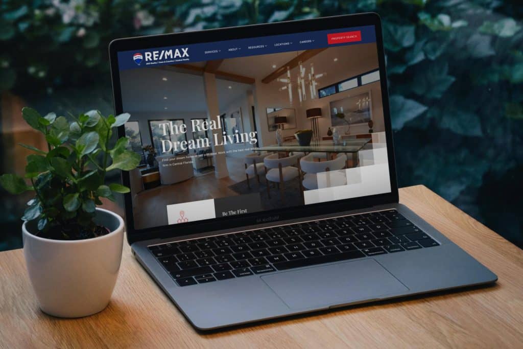 Laptop on a brown desk next to a houseplant. The laptop is showing the Re/Max website home page